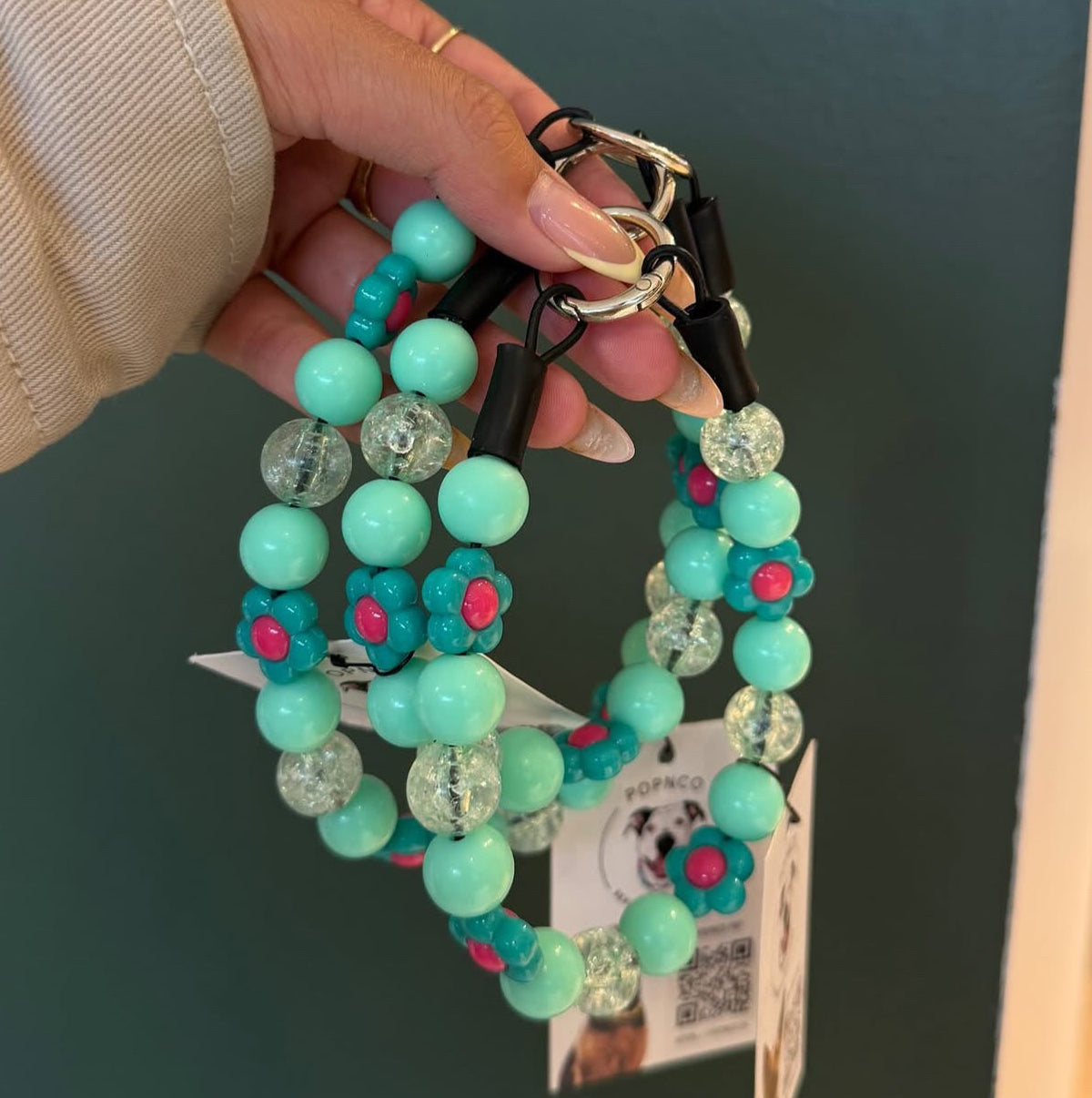 A hand wearing a cream-colored knit sweater holds a set of turquoise and teal beaded dog collars with black leather straps and gold-tone hardware. A pink text overlay at the top reads, 'I adore these new collars @popco_pnp just dropped off!'.