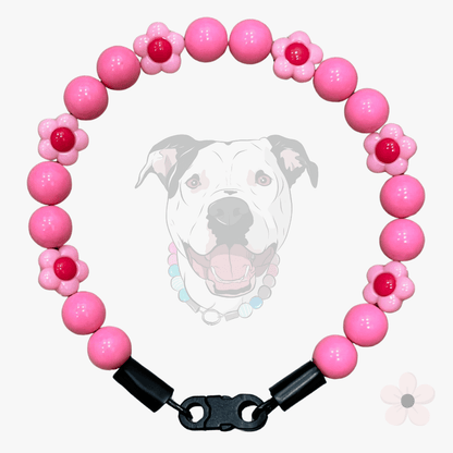 A pink beaded dog collar featuring round pink beads and flower-shaped beads with red centers, finished with black cylindrical spacer caps and a black lobster clasp, shown against a light gray background with a faint dog graphic.