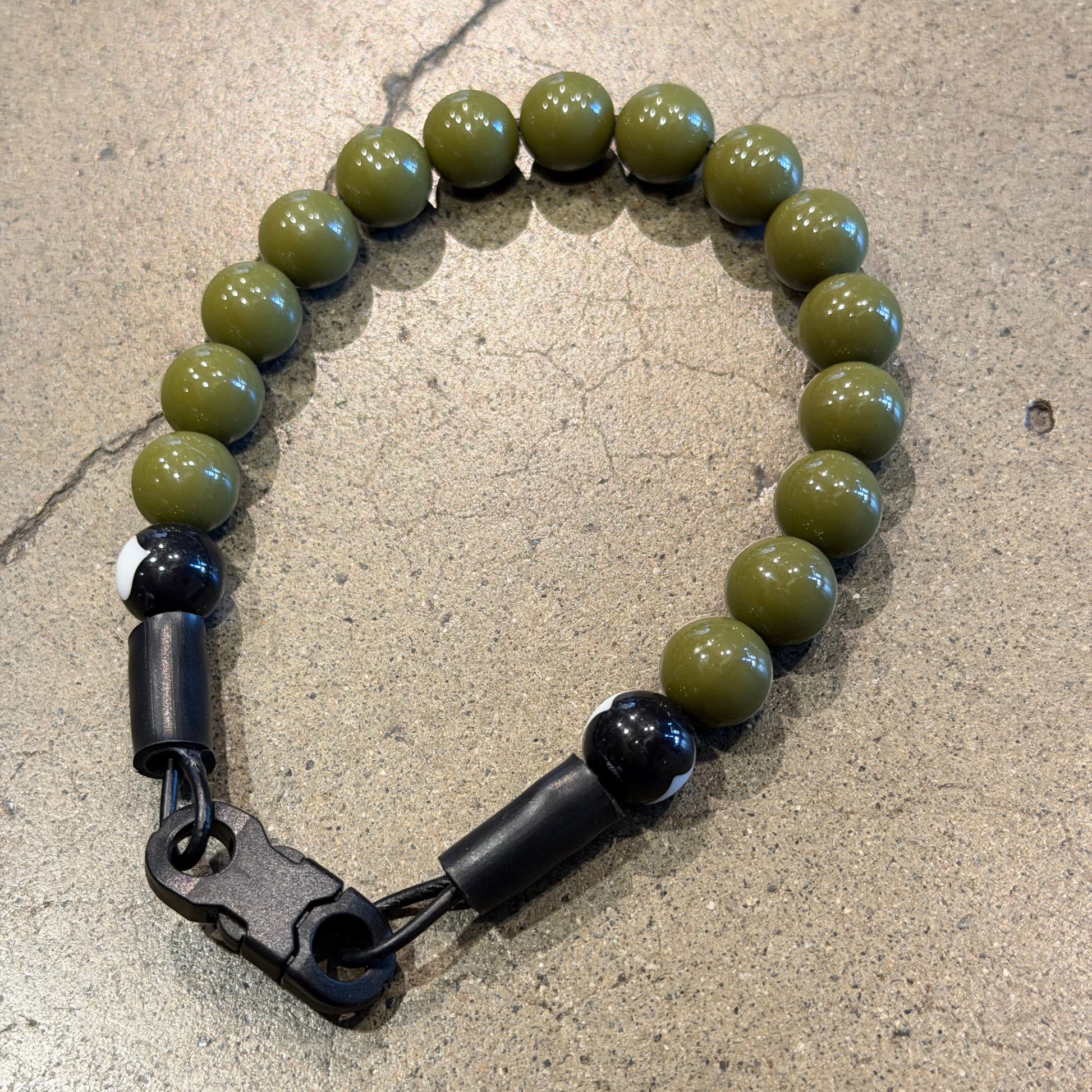 A beaded collar with olive green round beads, black spacer beads, and a black quick release buckle, displayed on a textured concrete floor.