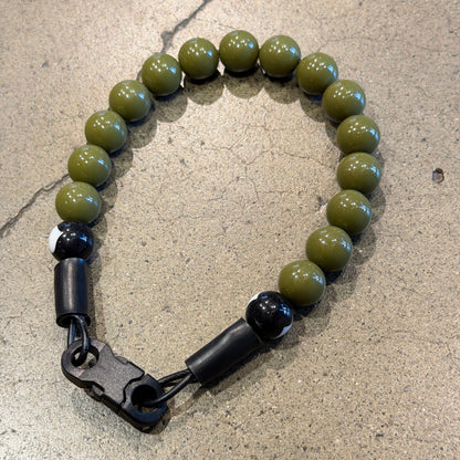 A beaded collar with olive green round beads, black spacer beads, and a black quick release buckle, displayed on a textured concrete floor.