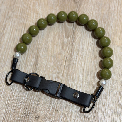 An olive green beaded bracelet with a black adjustable leather strap and metal hardware, displayed on a light wood grain surface.