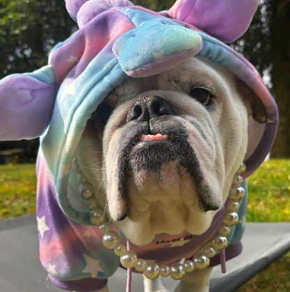 A bulldog wearing a pastel unicorn costume hoodie with a horn, ears, and pearl necklace, standing outdoors on a patio chair in a grassy yard.