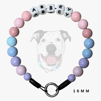 A pastel beaded dog collar with alternating pink, light blue, and lavender beads, featuring white letter beads spelling 'ABBEY' and a black clasp with a metal ring, shown against a light gray background with a faint dog graphic.
