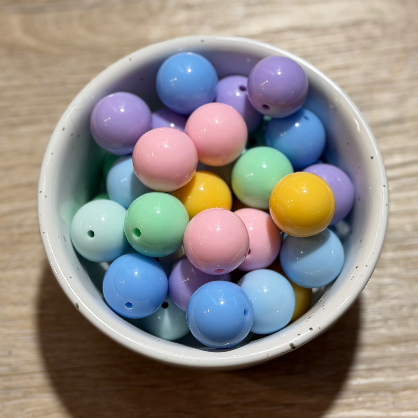 A small white ceramic bowl filled with pastel-colored wooden beads in shades of blue, pink, purple, green, and yellow, set on a light brown wooden tabletop.