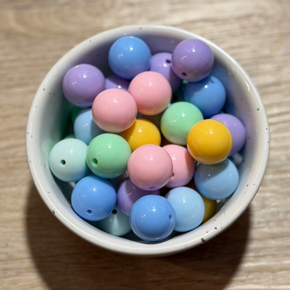 A small white ceramic bowl filled with pastel-colored wooden beads in shades of blue, pink, purple, green, and yellow, set on a light brown wooden tabletop.