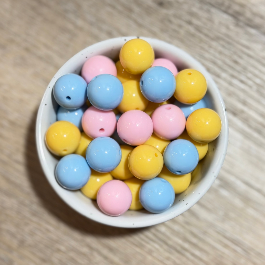 A top-down view of pastel-colored round beads in pink, blue, and yellow, arranged in a small white speckled bowl on a light wood surface.