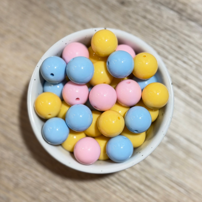 A top-down view of pastel-colored round beads in pink, blue, and yellow, arranged in a small white speckled bowl on a light wood surface.