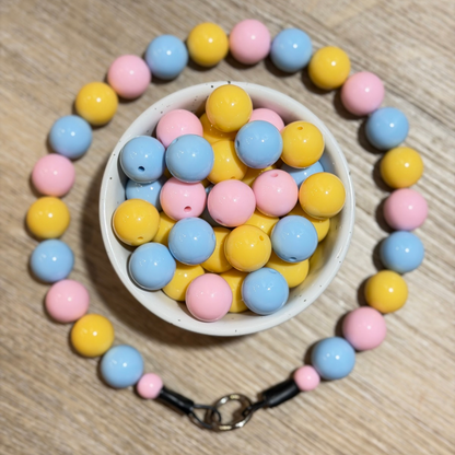 A pastel beaded pet collar made of pink, yellow, and light blue round beads, arranged around a small white bowl filled with matching beads on a light wood surface.