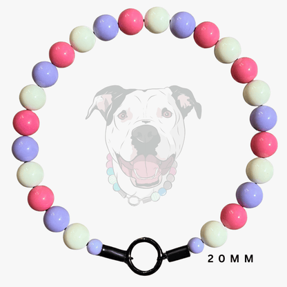 A beaded dog collar necklace made of glossy pastel beads in pink, lavender, cream, and light blue, arranged in a circle with a black toggle clasp. A faint dog face wearing the collar is visible in the background, and '20MM' is printed at the bottom right.