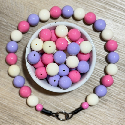 A beaded necklace made of pastel pink, lavender purple, and cream white round beads, arranged in a circle around a small white bowl filled with loose matching beads. The necklace has a black cord and a silver metal clasp.