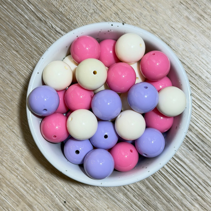 A bowl filled with round craft beads in pastel pink, lavender purple, and off-white, arranged on a light wood surface.