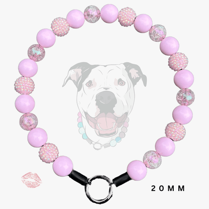 A pink beaded dog collar with alternating smooth light pink beads and textured pink-and-white speckled beads, featuring a silver metal ring clasp and black connector ends. A faint dog face graphic appears behind the collar, with a small lip print and '20MM' text on a light gray background.
