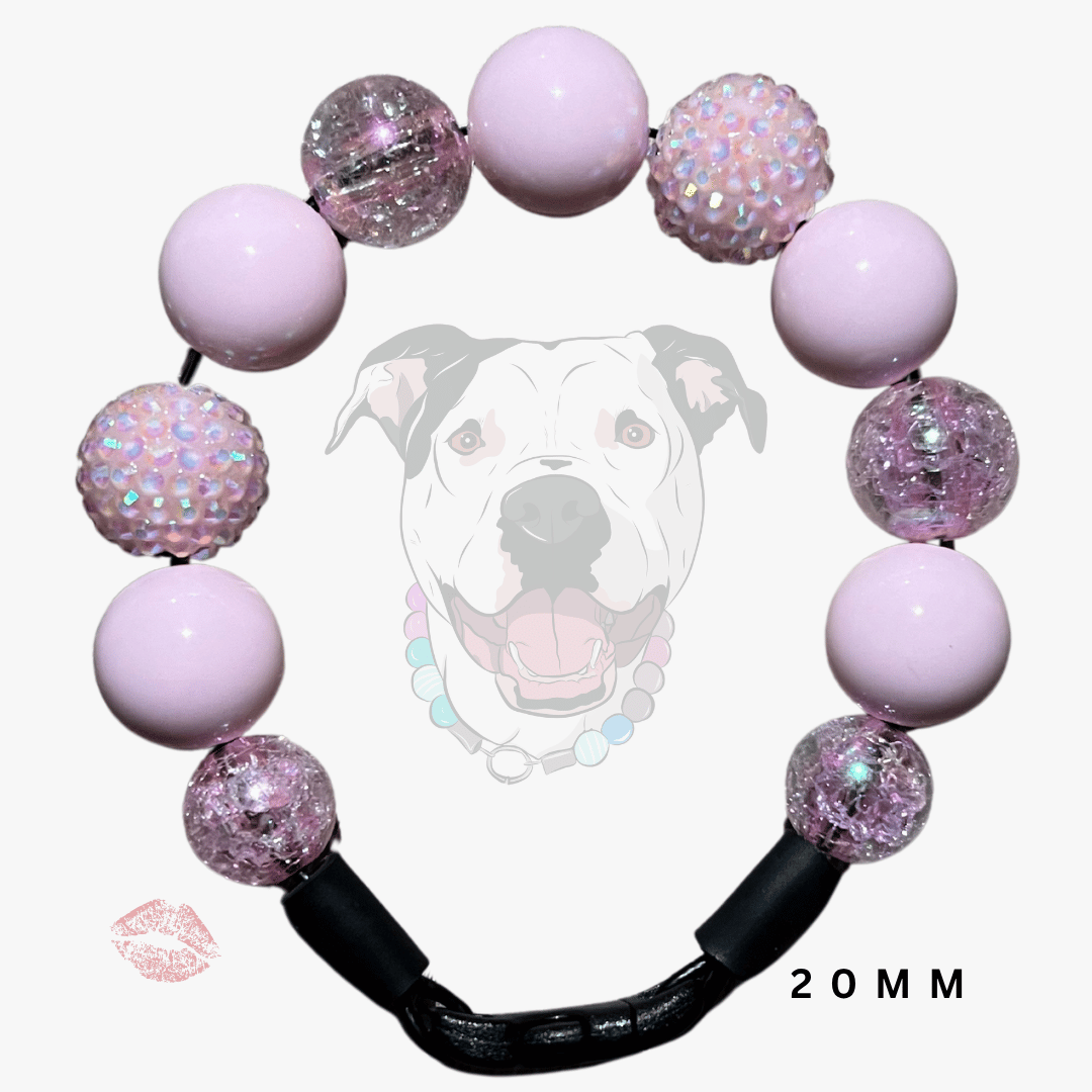 A pink beaded dog collar or bracelet-style accessory with light pink round beads, some clear glitter-filled beads, and textured iridescent beads, plus a black clasp. A faint dog face graphic and a small lip mark appear behind and near the product on a light gray background, with '20MM' text at the bottom right.