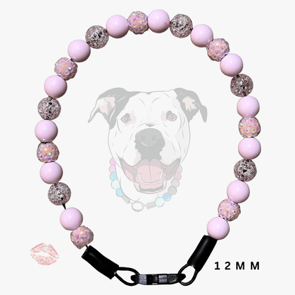 A pink dog collar made of alternating smooth light pink beads and glittery pink-and-white patterned beads, with a black metal clasp and black end caps. A faint dog face graphic appears behind the collar, along with a small lip mark and the text '12 MM'.