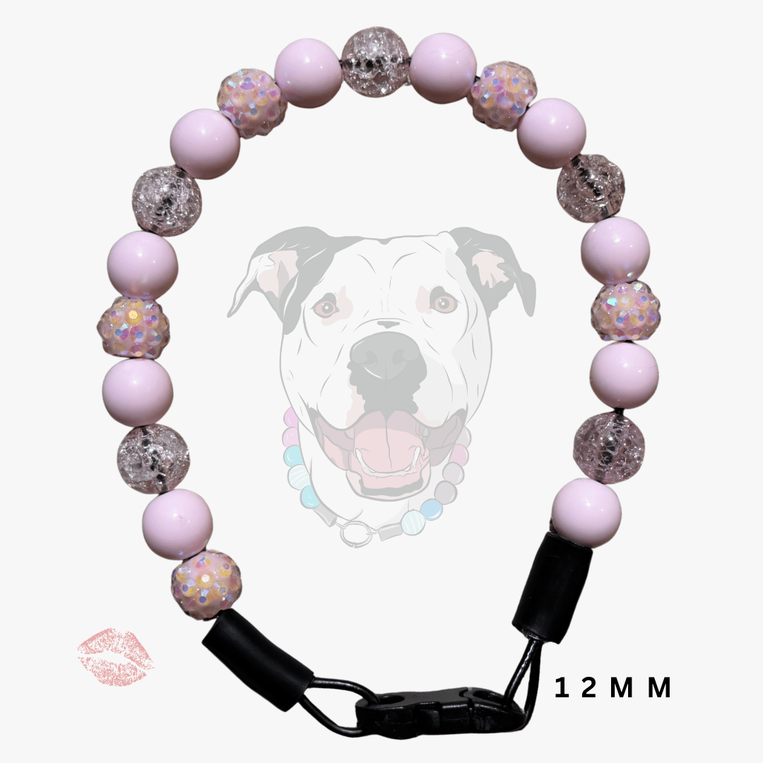 A beaded dog collar with light pink and glittery silver beads, featuring a black plastic buckle and clip closure. A faint graphic of a dog's face is visible in the background, along with a small pink lip print and '12 MM' text.