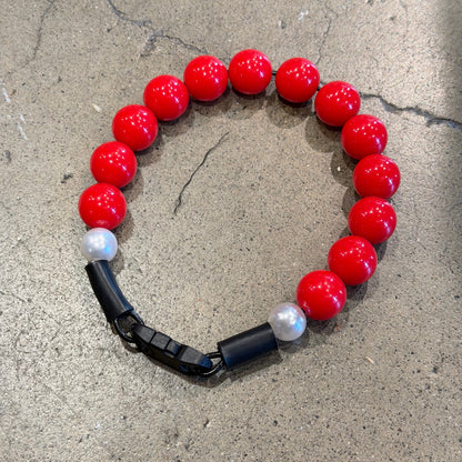 A beaded bracelet with glossy red round beads, two small white pearl-like beads, and black metal connector pieces, photographed on a textured gray concrete surface.