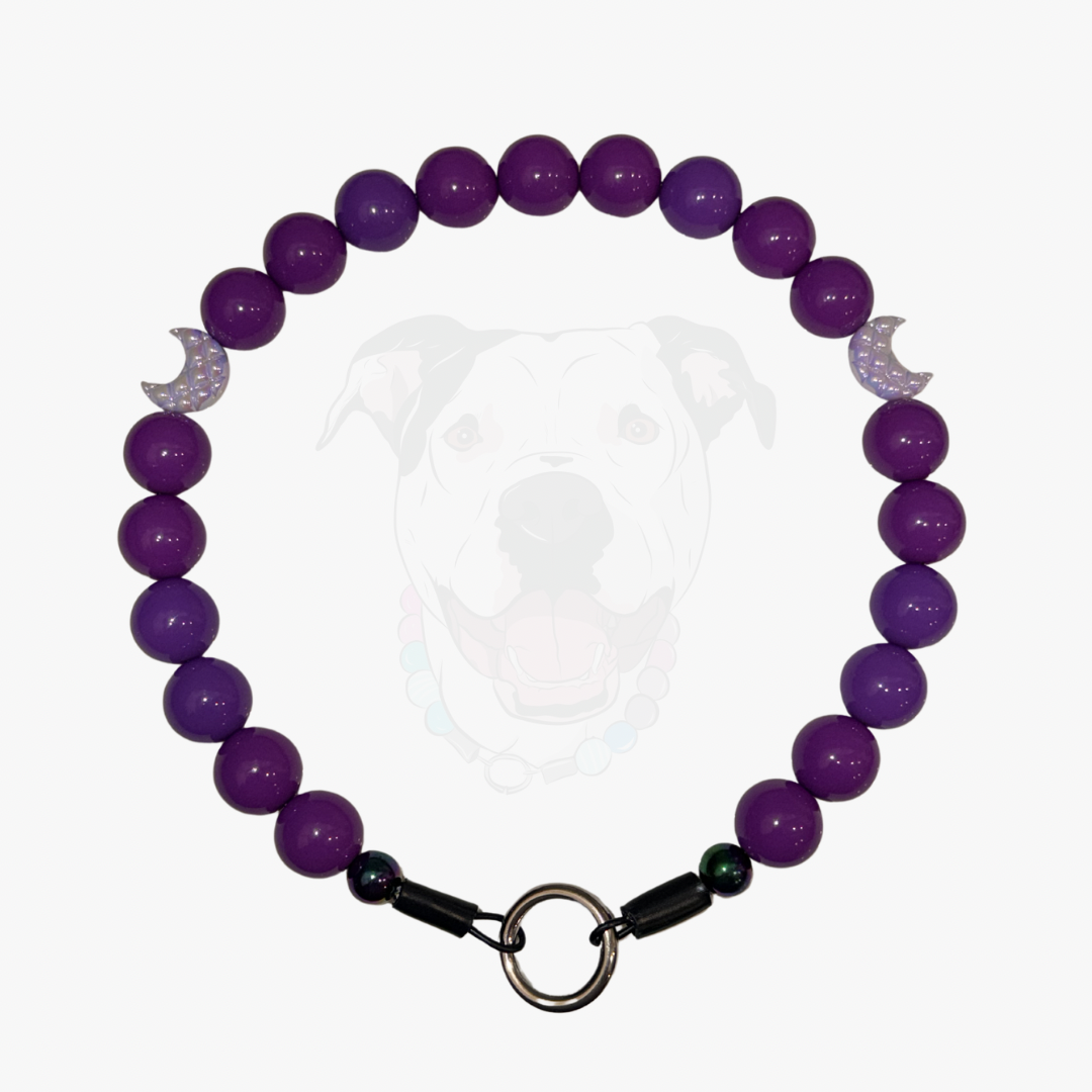A purple beaded pet collar featuring glossy round purple beads, a silver crescent moon charm with a clear rhinestone center, black spacer beads, and a silver metal ring clasp, shown against a light gray background with a faint dog graphic.