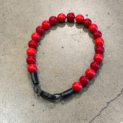 A beaded bracelet made of glossy red round beads with a black metal clasp, photographed on a textured gray concrete surface.