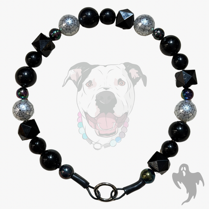 A beaded dog collar featuring glossy black round beads, spiderweb-patterned silver beads, and faceted black geometric beads, displayed against a light gray background with a faint dog graphic.