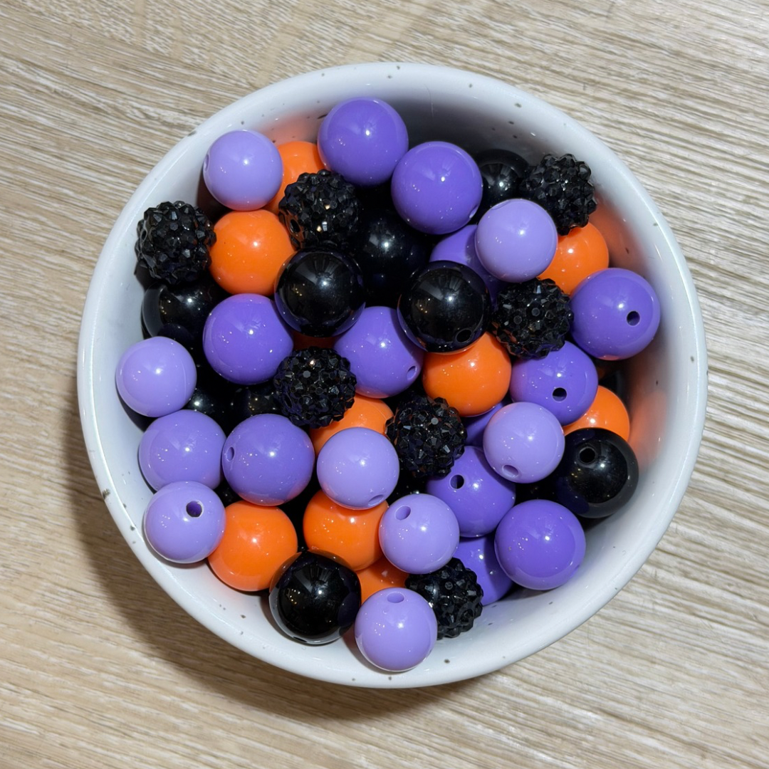 A white speckled bowl filled with mixed craft beads, including purple, orange, black, and glittery black beads, on a light wood surface.