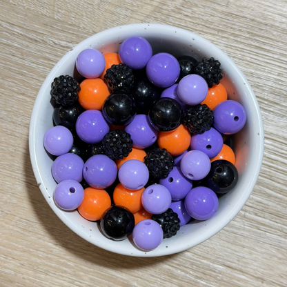 A white speckled bowl filled with mixed craft beads, including purple, orange, black, and glittery black beads, on a light wood surface.
