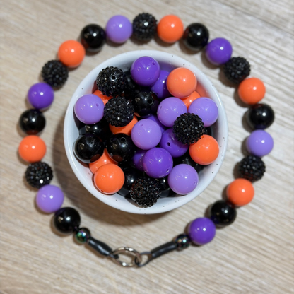 A beaded bracelet arranged in a circle around a small white bowl filled with loose beads. The bracelet features glossy black, matte orange, and lavender-purple round beads, with a silver clasp visible at the bottom.