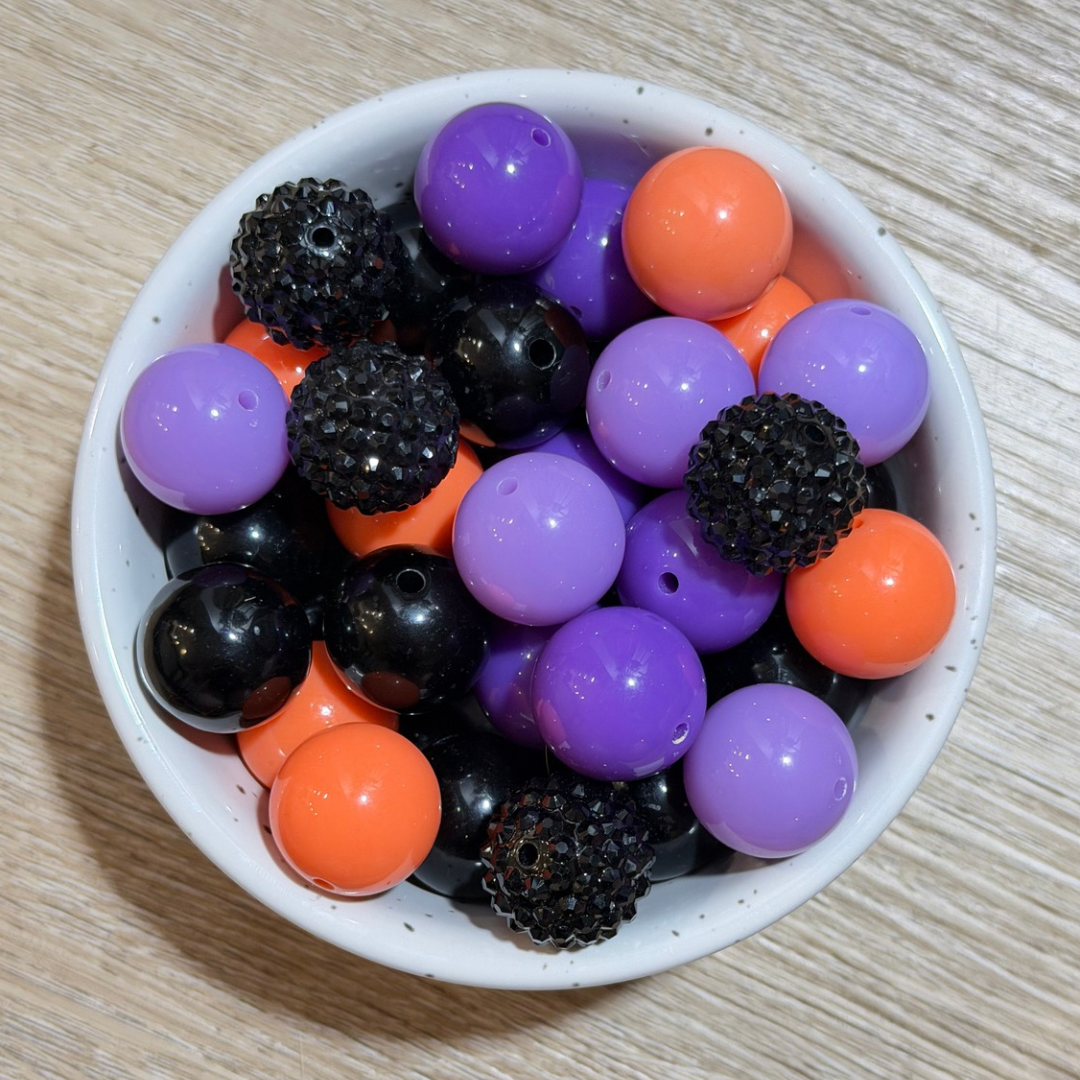 A white ceramic bowl filled with craft beads in purple, orange, and black, photographed from above on a light wood surface.