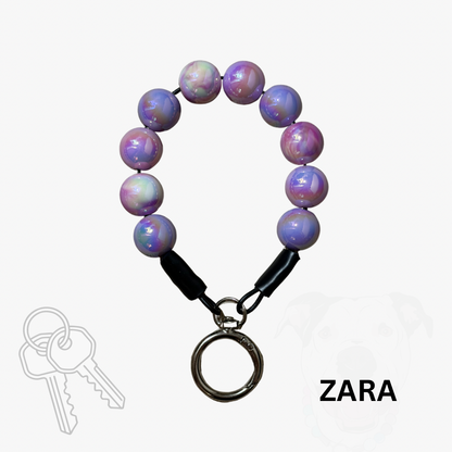 A beaded keychain with iridescent purple, pink, and blue beads, a black loop connector, and a silver metal key ring clasp, shown on a light gray background with a 'ZARA' text overlay and a faint key graphic.