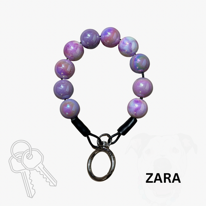 A beaded keychain bracelet with iridescent purple and pink beads, black connector caps, and a silver metal key ring, shown on a light gray background with a key graphic and the text 'ZARA'.