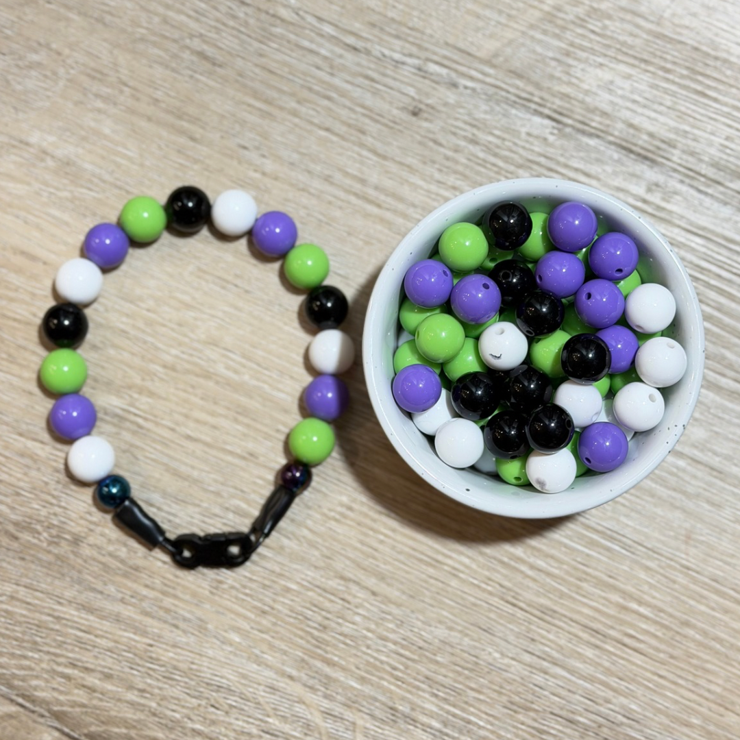 A beaded pet collar with purple, green, black, and white beads is displayed next to a small white bowl filled with matching loose beads on a light wood surface.