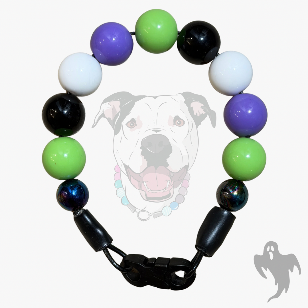 A beaded dog collar featuring alternating solid purple, green, white, and black round beads, with two iridescent blue beads near the clasp. The collar is displayed against a light gray background with a faint graphic of a pitbull dog wearing the collar and a small ghost icon in the bottom right corner.