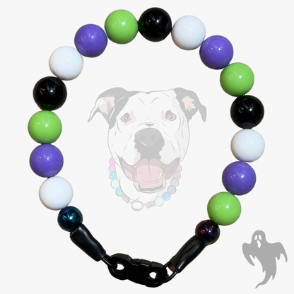 A beaded dog collar with alternating glossy beads in purple, green, black, white, and one iridescent blue bead, separated by black spacer beads and a black clasp. A faint dog face graphic is visible in the background.