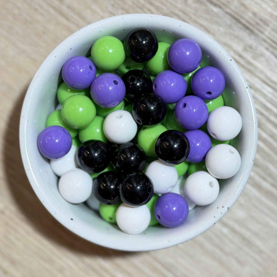 A white ceramic bowl filled with glossy round beads in purple, lime green, black, and white, arranged on a light wood surface.