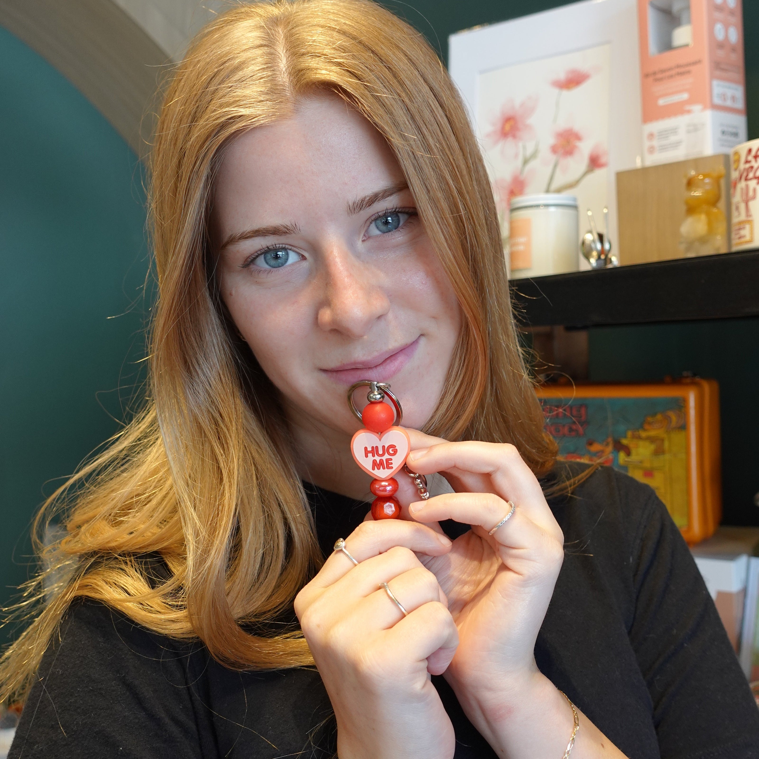 A young woman with blonde hair holds a small red and white heart-shaped keychain or charm in front of her face, wearing a black t-shirt in an indoor setting with shelves in the background.