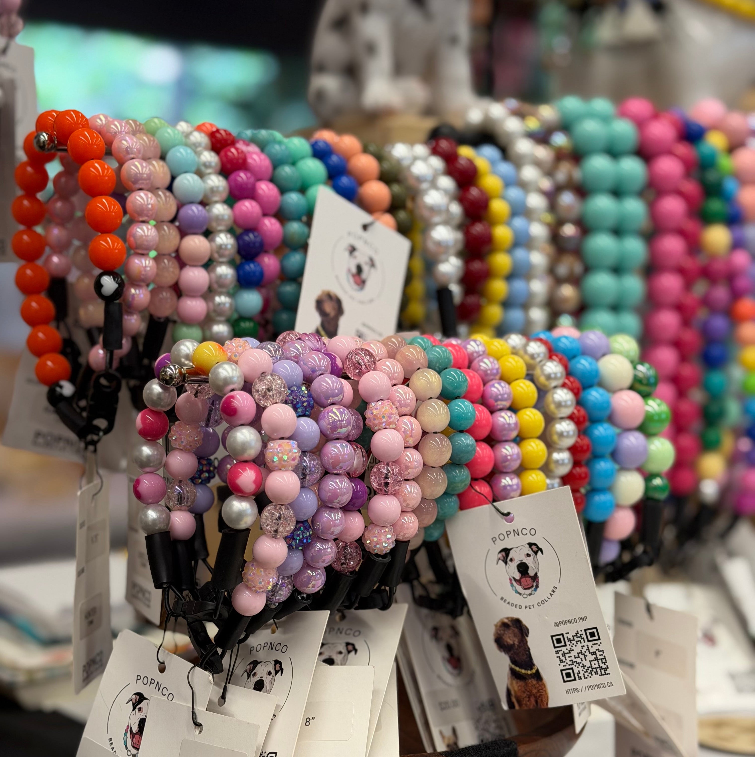 A display of colorful beaded dog collars and bracelets from the brand POPNCO, featuring various pastel and bright colors on a tabletop with product tags attached.