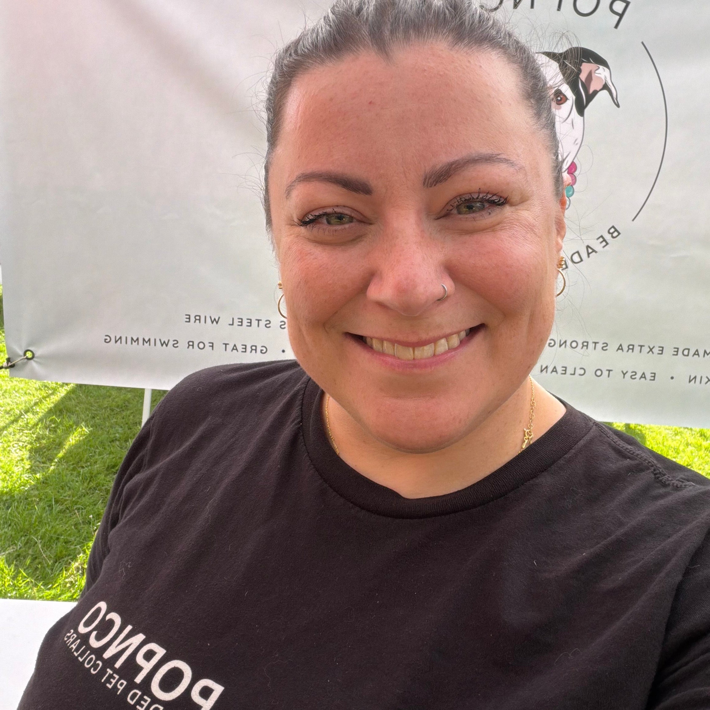 A smiling woman wearing a black t-shirt with white 'Popnco beaded pet collars' branding, standing outdoors in front of a white banner.