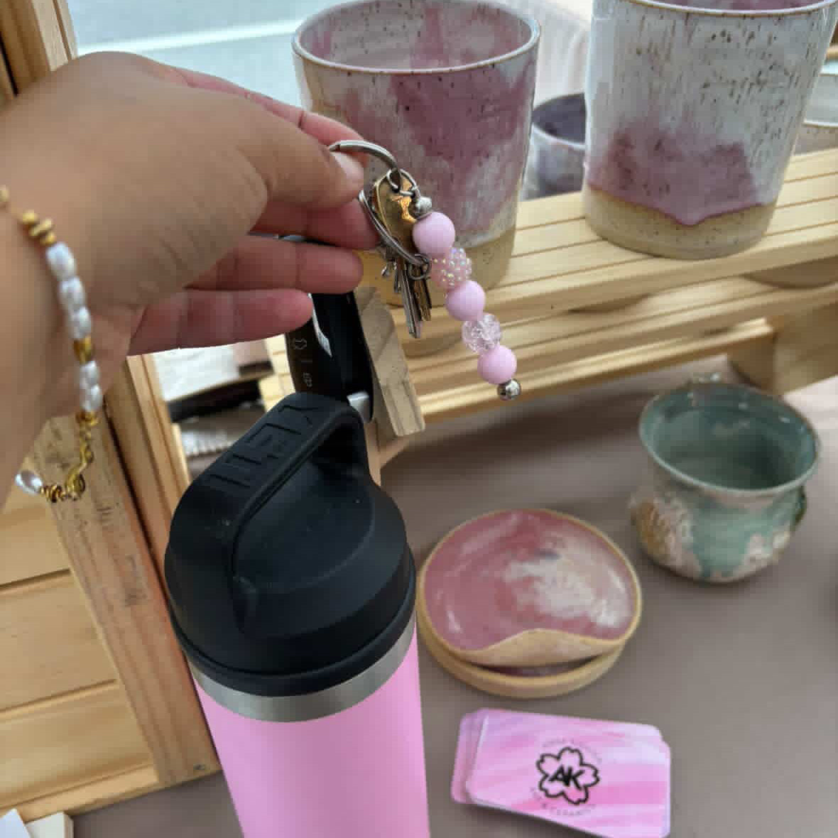 A pink insulated tumbler with a black lid is shown in a market or shop setting, with a hand holding a keychain featuring pink beads. Ceramic cups and a small pink item are visible on a wooden display stand in the background.