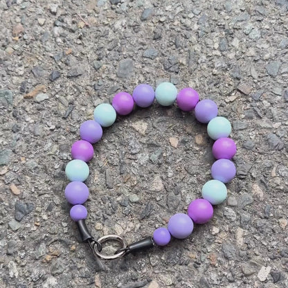 A beaded pet collar with alternating light purple, lavender, and pale mint-green beads, featuring an O ring slip on style and made on Aircraft Grade Stainless Steel Wire laying Flat on a concrete background 